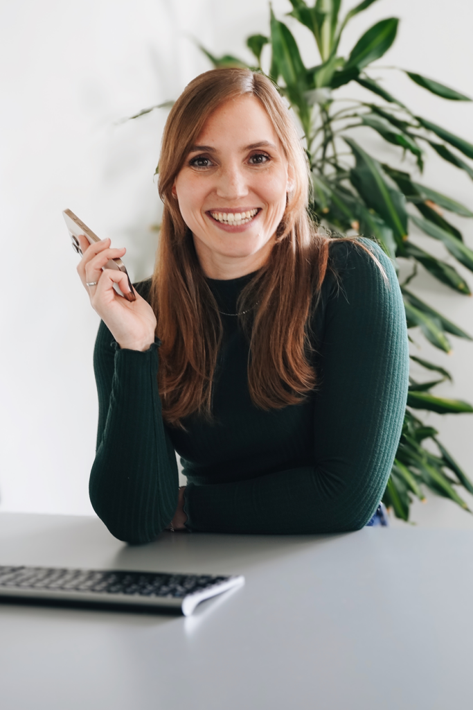 Die Gründerin Lisa Müller trägt einen grünen Pullover und sitzt an ihrem Schreibtisch mit dem Handy in der Hand.
